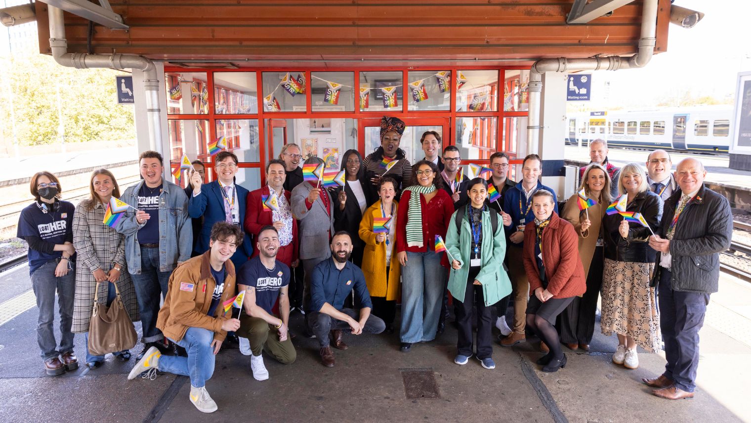 A large group of people standing or kneeling outside a waiting room on a station platform