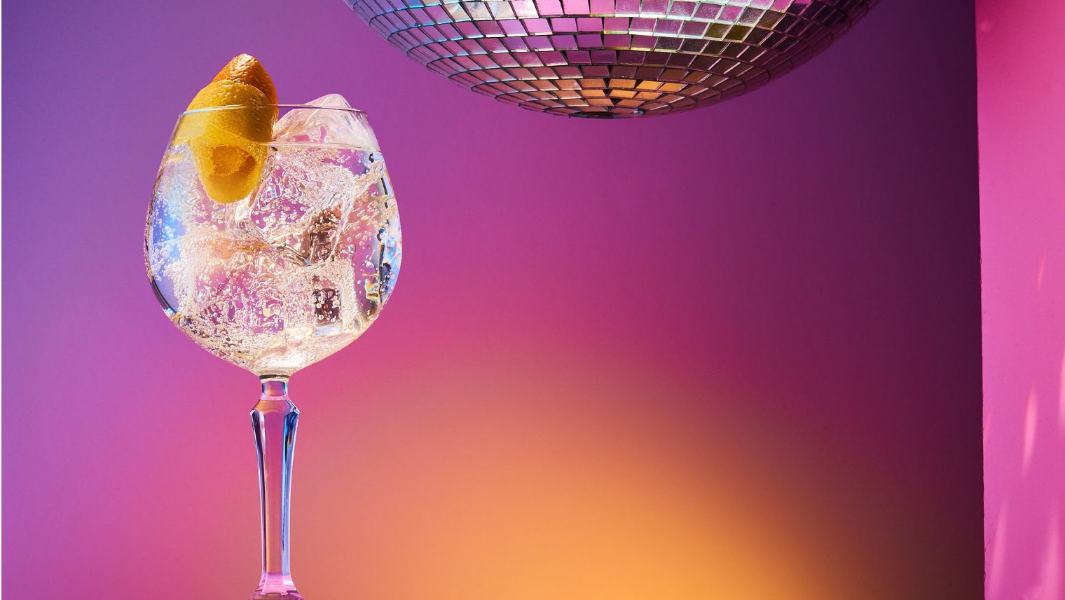 A cocktail with ice in a wine glass in front of a pink and orange background and glitter ball above