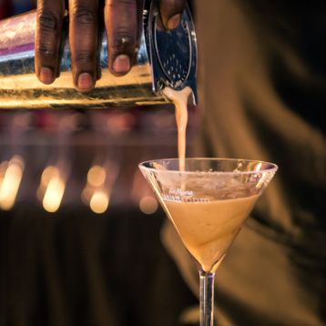 A hand holding a cocktail shaker pours a liquid into a cocktail glass