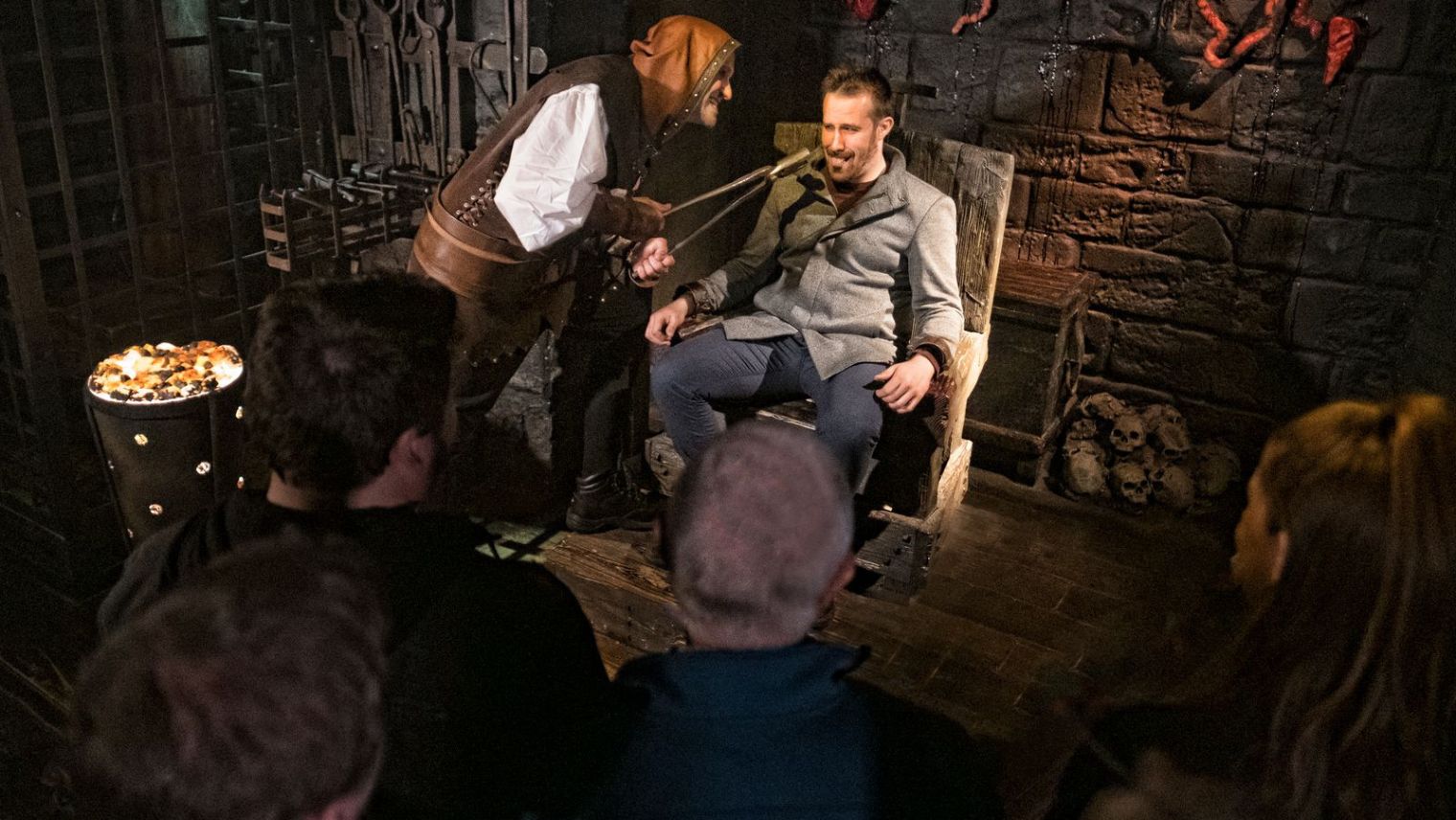 A character in a gloomy lit room in The London Dungeon  leans menacingly towards a man dressed in modern clothes who is seated in a chair with his hands strapped to the arms. The character in old Victorian costume is hold a pair of large plyers to the man's mouth while onlookers watch